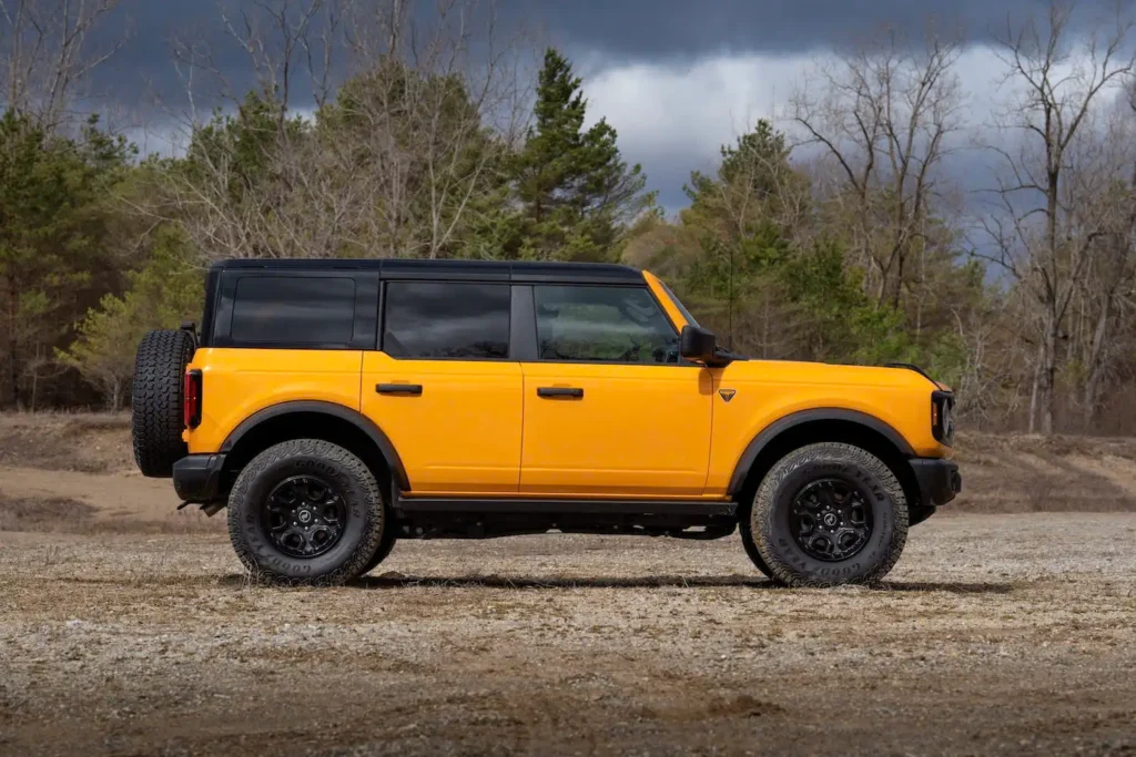 Side profile of an orange Ford Bronco parked outdoors, featuring a black contrast roof, full-size rear-mounted spare tire, off-road tires, and boxy SUV proportions against a wooded landscape under overcast skies