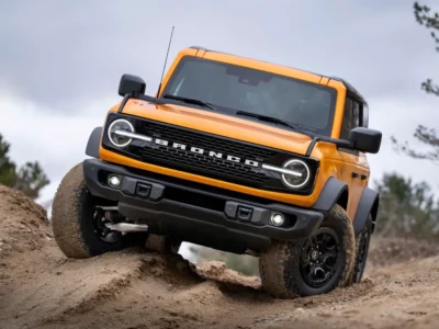 Orange Ford Bronco navigating a rugged off-road terrain with visible suspension articulation, chunky all-terrain tires, signature round headlights, black fender flares, and a steep dirt obstacle in an outdoor trail setting