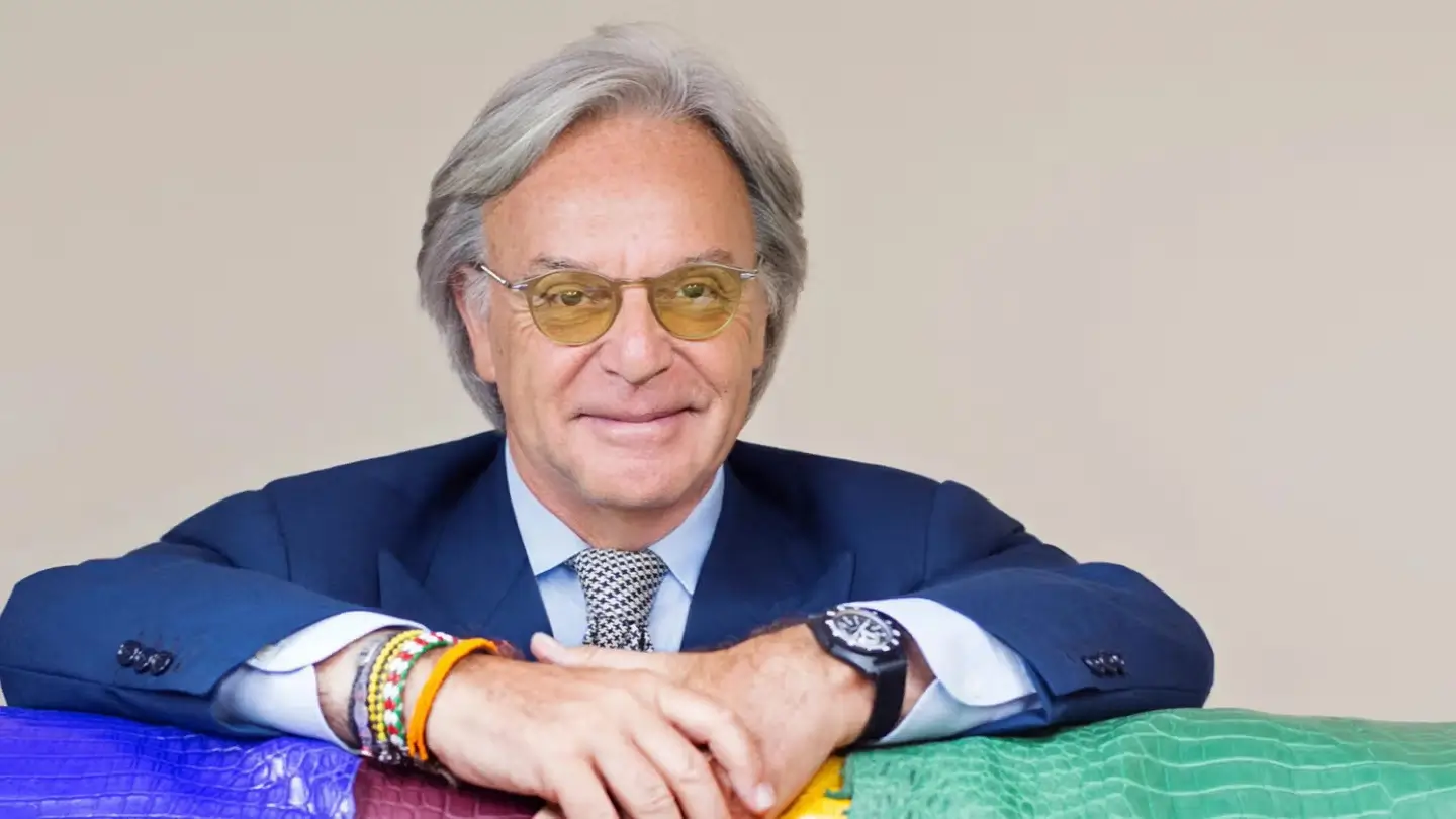 Portrait of Diego Della Valle wearing a tailored navy suit, patterned tie, and lightly tinted glasses while leaning across colorful leather surfaces, reflecting the refined luxury aesthetic and artisanal identity closely associated with Tod’s Group