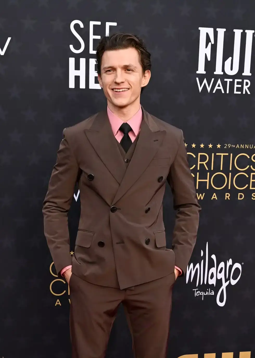 A polished red-carpet portrait of a young actor wearing a sharply tailored chocolate-brown double-breasted suit paired with a soft pink dress shirt and slim black tie. The structured tailoring, satin-like sheen of the fabric, and layered waistcoat styling give the look a refined yet contemporary formalwear aesthetic. Standing confidently with hands in pockets, the actor smiles against a dark awards-show backdrop featuring sponsor logos and gold typography, creating a clean Hollywood premiere atmosphere that balances classic menswear elegance with youthful modern styling
