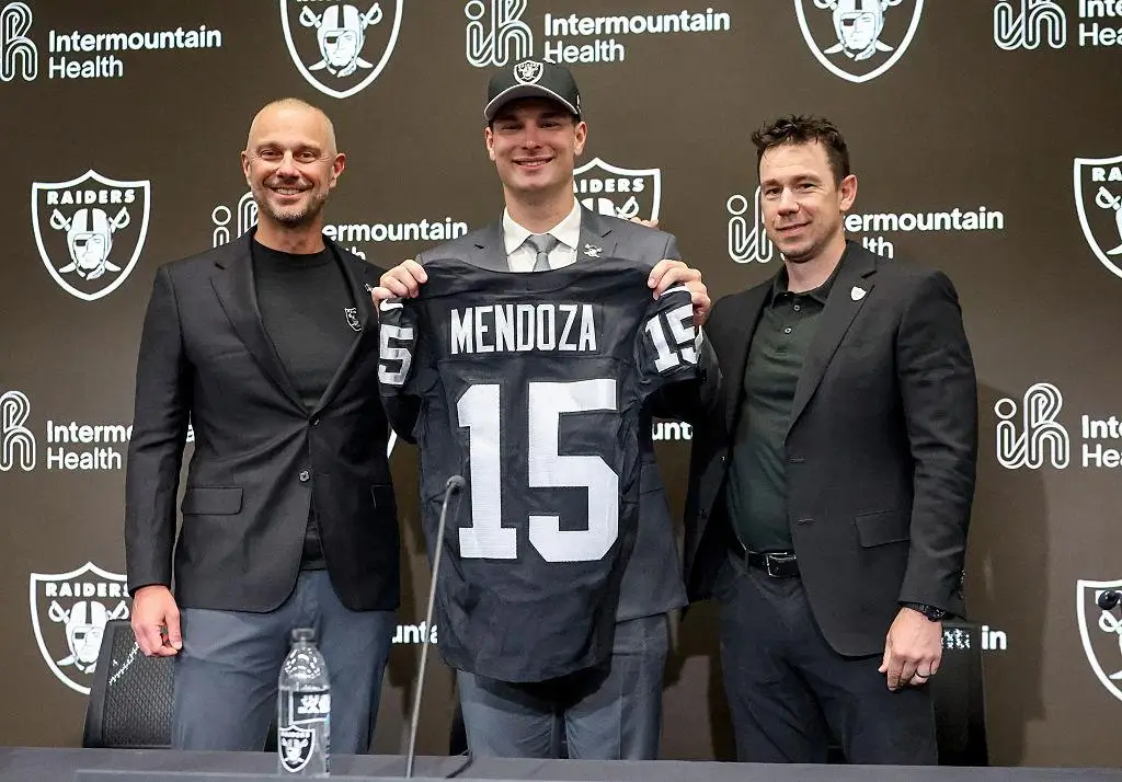 Three men stand at a press conference backdrop with Las Vegas Raiders logos, as a young player in a suit and Raiders cap holds up a black jersey reading “MENDOZA 15,” flanked by two team representatives in dark suits
