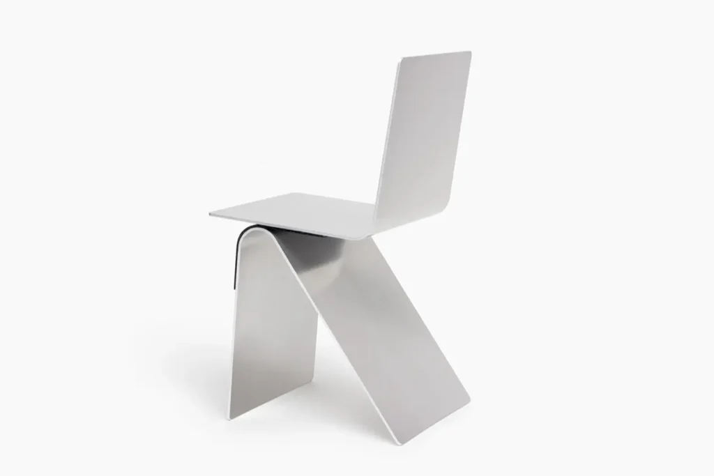 Full product shot of a sculptural minimalist metal chair crafted from folded brushed aluminum, featuring sharp geometric lines, curved structural bends, and an industrial monochrome aesthetic against a clean studio backdrop