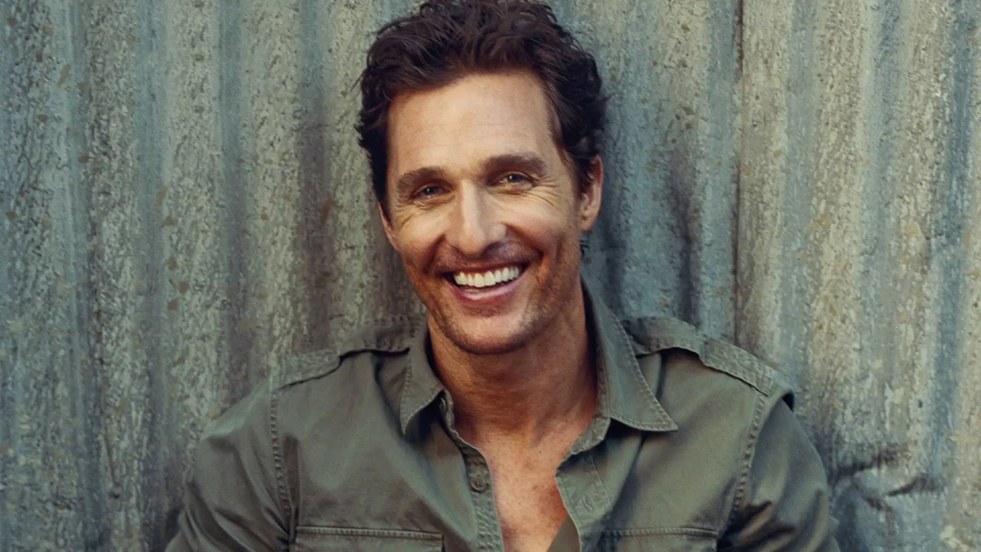 Portrait of actor Matthew McConaughey smiling against a weathered textured wall while wearing an olive green button-up shirt in a relaxed outdoor-style editorial photograph