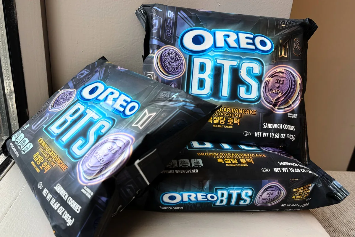 Multiple packs of the limited-edition Oreo x BTS sandwich cookies displayed on a beige surface near a window. The packaging features glowing blue “OREO BTS” branding, purple cookie graphics, and brown sugar pancake flavor labeling inspired by Korean hotteok