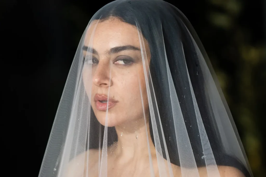 Close-up portrait of a woman beneath a sheer translucent bridal veil, featuring dark hair, smoky eye makeup, and soft lighting that creates a cinematic gothic editorial atmosphere
