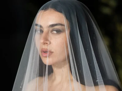 Close-up portrait of a woman beneath a sheer translucent bridal veil, featuring dark hair, smoky eye makeup, and soft lighting that creates a cinematic gothic editorial atmosphere