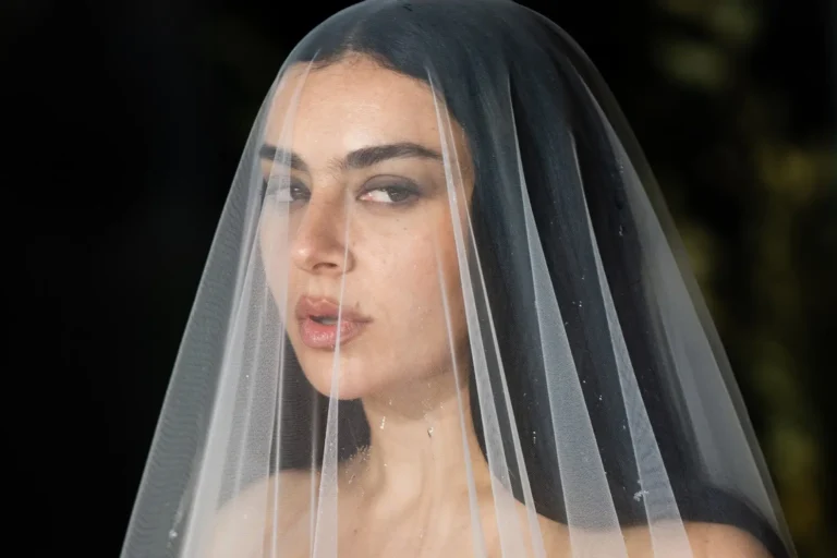Close-up portrait of a woman beneath a sheer translucent bridal veil, featuring dark hair, smoky eye makeup, and soft lighting that creates a cinematic gothic editorial atmosphere