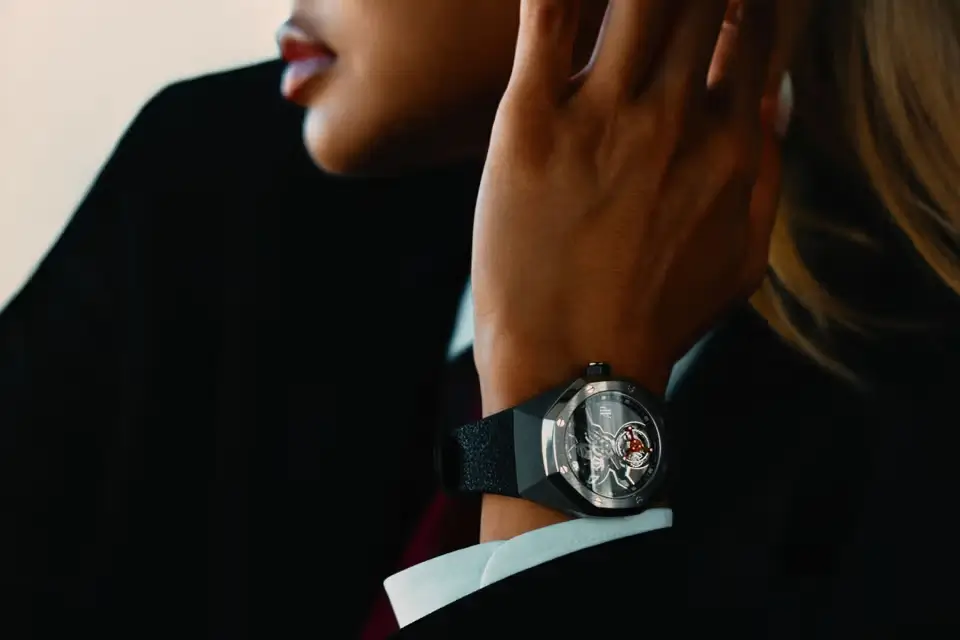 Editorial close-up of a model in dark tailored styling wearing a titanium-toned luxury watch with an openworked dial and prominent red flying tourbillon detail. The wrist is positioned near the face, emphasizing the architectural case shape, textured black strap, and refined high-fashion campaign aesthetic