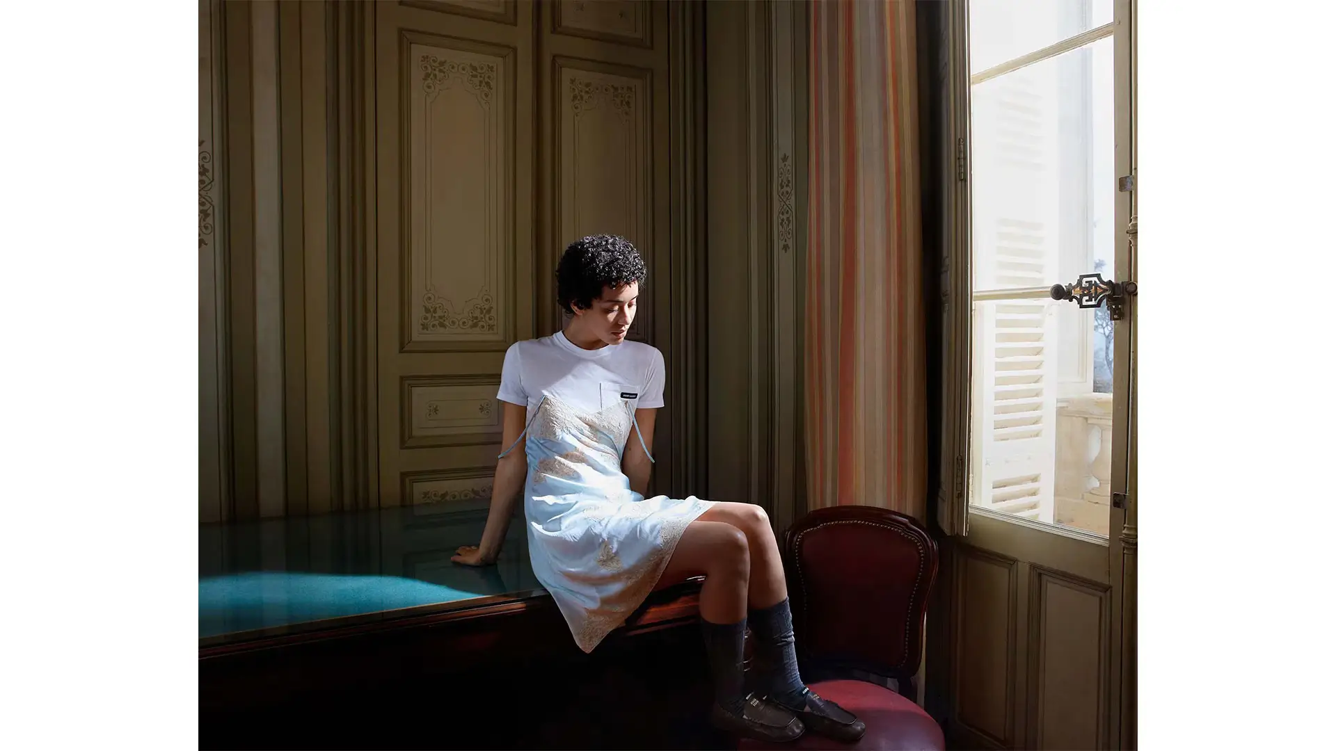 Young woman seated on a dark table inside an elegant sunlit room, wearing a white layered slip dress with knee-high socks and loafers beside an open window with vintage architectural detailing
