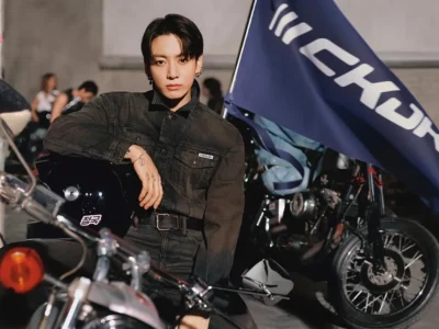 Editorial campaign portrait of a young man seated among motorcycles wearing a dark washed denim set with subtle branded detailing. He faces the camera while resting one arm on a glossy black helmet, with a large CKJK flag and parked bikes creating a moody garage backdrop that reinforces the collection’s biker-inspired aesthetic