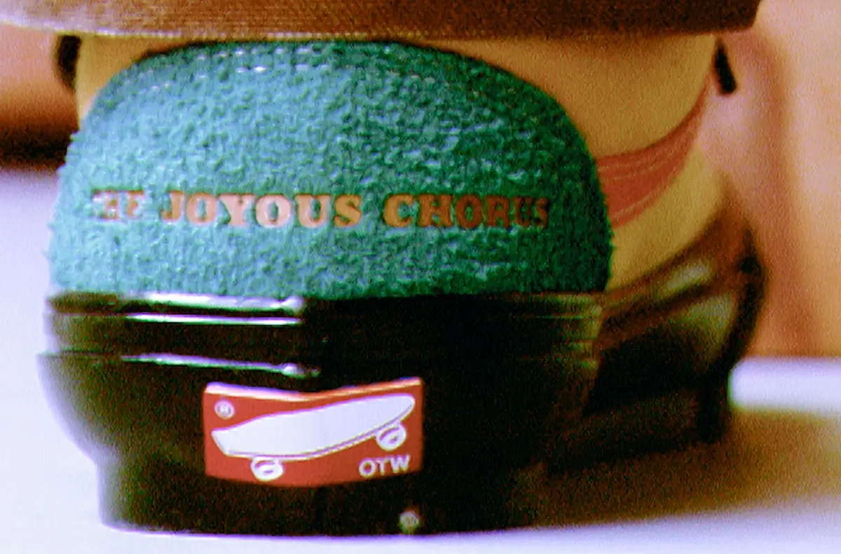 Close-up rear view of an OTW by Vans shoe featuring textured teal suede with gold “The Joyous Chorus” heel branding, a glossy black midsole, and a red OTW skateboard logo heel tab captured in a grainy cinematic aesthetic
