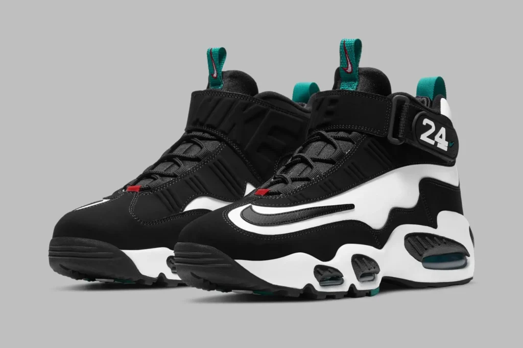 Angled front-side view of a pair of black-and-white Nike Griffey-inspired high-top sneakers featuring oversized outlined Swooshes, sculpted wave-like paneling, and chunky midsoles with visible teal-tinted Air cushioning units. The right shoe displays a velcro ankle strap stamped with the number “24,” while teal pull tabs and small red lace accents add contrast throughout the design. The shoe are photographed against a smooth gray studio background, emphasizing their bold late-1990s baseball aesthetic