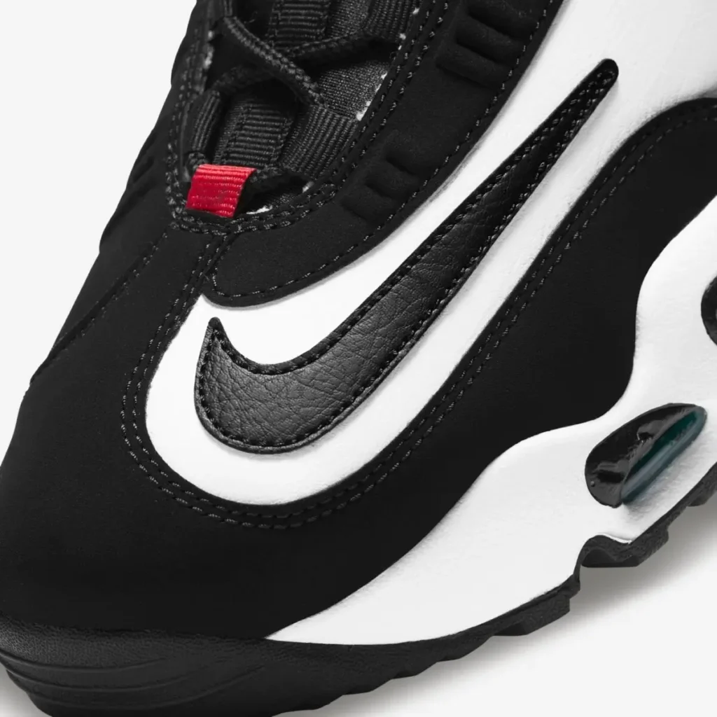 Close-up detail of a black-and-white Nike Griffey-inspired shoe highlighting the oversized layered Swoosh design along the side panel. The image showcases black nubuck-like materials, textured leather Swoosh overlays, contrast stitching, and a sculpted white midsole with a view teal-tinted Air cushioning unit. A small red lace loop accent near the forefoot adds a subtle pop of color against the monochromatic upper