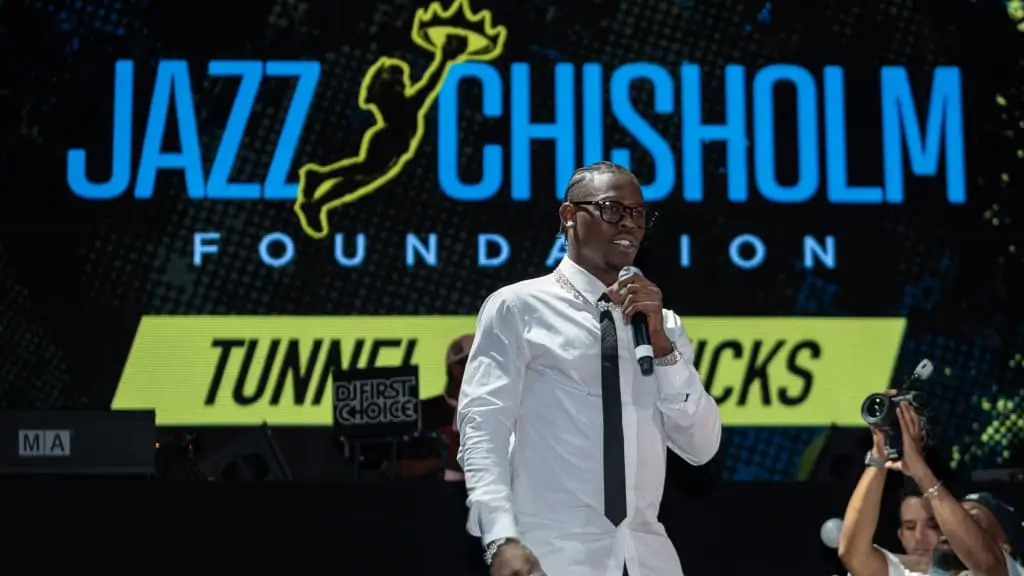 Jazz Chisholm Jr. poses with friends during a relaxed nighttime gathering, wearing a graphic oversized shirt, black trousers, luxury watch, and tinted glasses while seated against a dark modern interior backdrop
