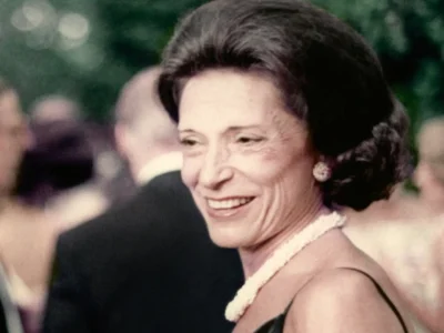 A softly lit archival portrait of Doris Fisher smiling during a formal social gathering, wearing pearl jewelry and an elegant black dress while surrounded by blurred guests in the background. The candid image captures her understated sophistication and warm presence, reflecting the quiet strength and refined sensibility that helped shape Gap Inc. and the broader cultural identity of San Francisco