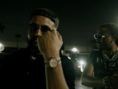 NAV and Quavo stand beside luxury vehicles under dim nighttime lighting in Miami during the “MUTT” music video, wearing dark clothing, sunglasses, layered jewelry, and watches against a shadowy street backdrop lined with palm trees and headlights
