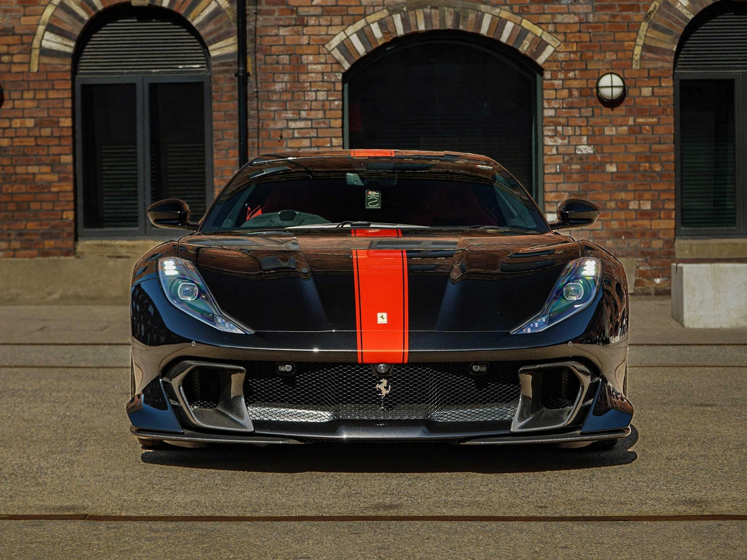 Front view of a black Ferrari 458 with a bold red racing stripe, parked on a paved street, featuring sharp headlights, large air intakes, and a low aggressive stance against a brick building with arched windows