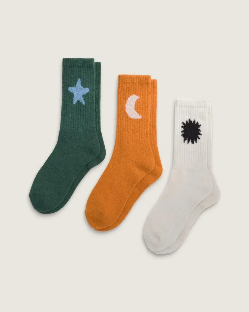 Three pairs of ribbed crew socks displayed against a neutral backdrop, featuring playful graphic motifs including a light blue star on dark green, a white crescent moon on orange, and a black abstract sunburst on white