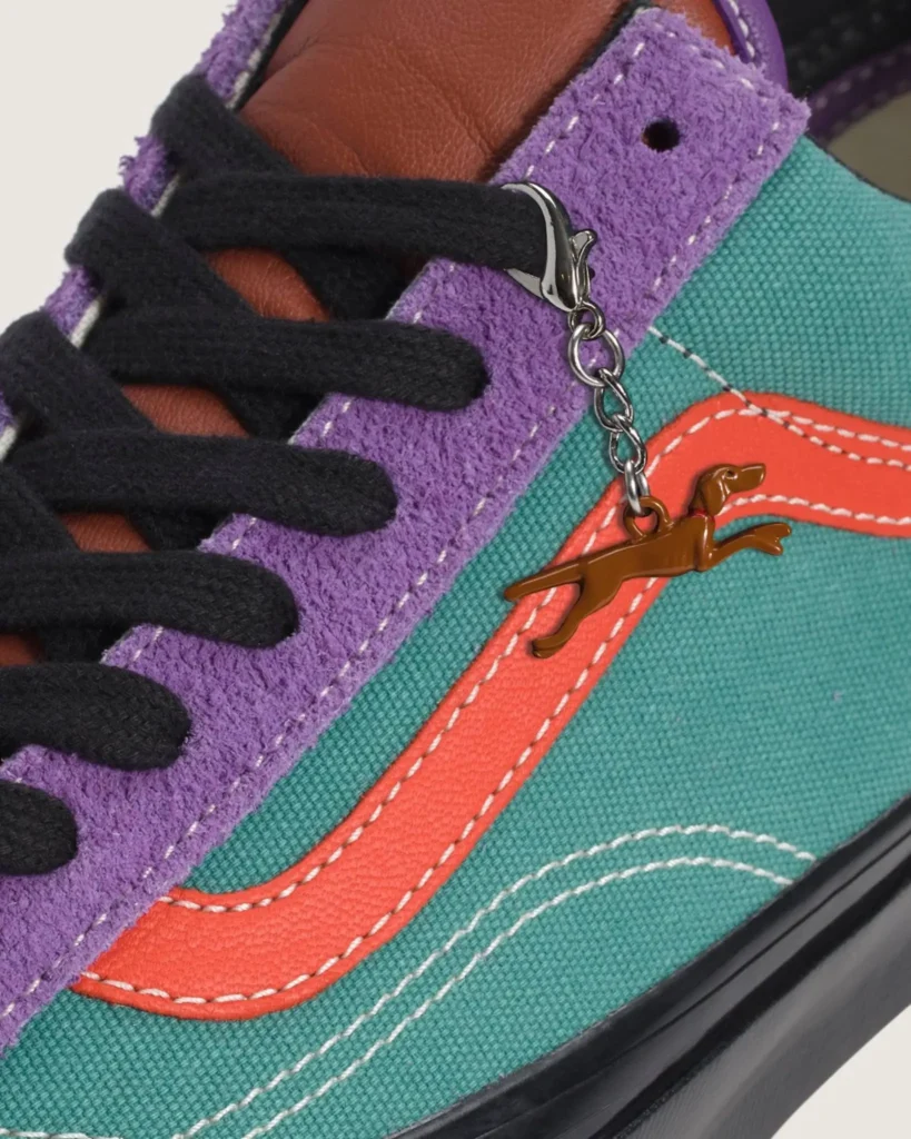Close-up detail of an OTW by Vans shoe featuring teal canvas panels, lavender hairy suede overlays, coral side stripe accents, black laces, and a dangling brown dog-shaped metallic charm attached near the eyelets, emphasizing the shoe’s textured handcrafted finish