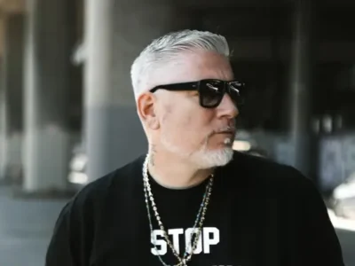 Portrait of musician Everlast standing outdoors beneath an urban overpass, wearing black sunglasses, layered chain necklaces, and a black graphic T-shirt. His silver hair is slicked back, and he gazes to the side against a softly blurred industrial city backdrop