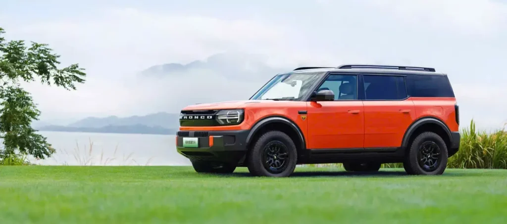 Orange Ford Bronco SUV shown in side profile on a grassy lakeside landscape, featuring a black contrast roof, rugged wheels, upright proportions, and an adventure-oriented design with mountains in the background