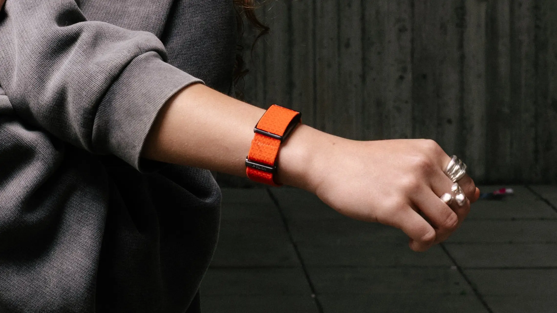 Close-up editorial shot of the bright orange Luna Band worn with layered silver rings and an oversized grey sweatshirt, emphasizing the textured strap and compact display-free wear profile