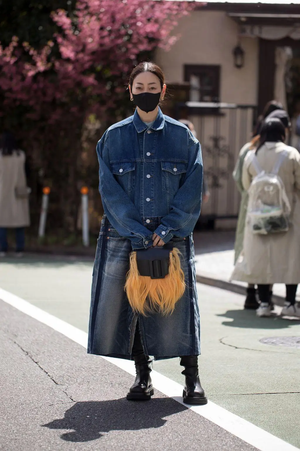 Street style portrait of a woman wearing an oversized indigo denim Type III jacket and long skirt, styled with black boots and a statement fur pouch, standing on a quiet residential street with cherry blossoms in bloom