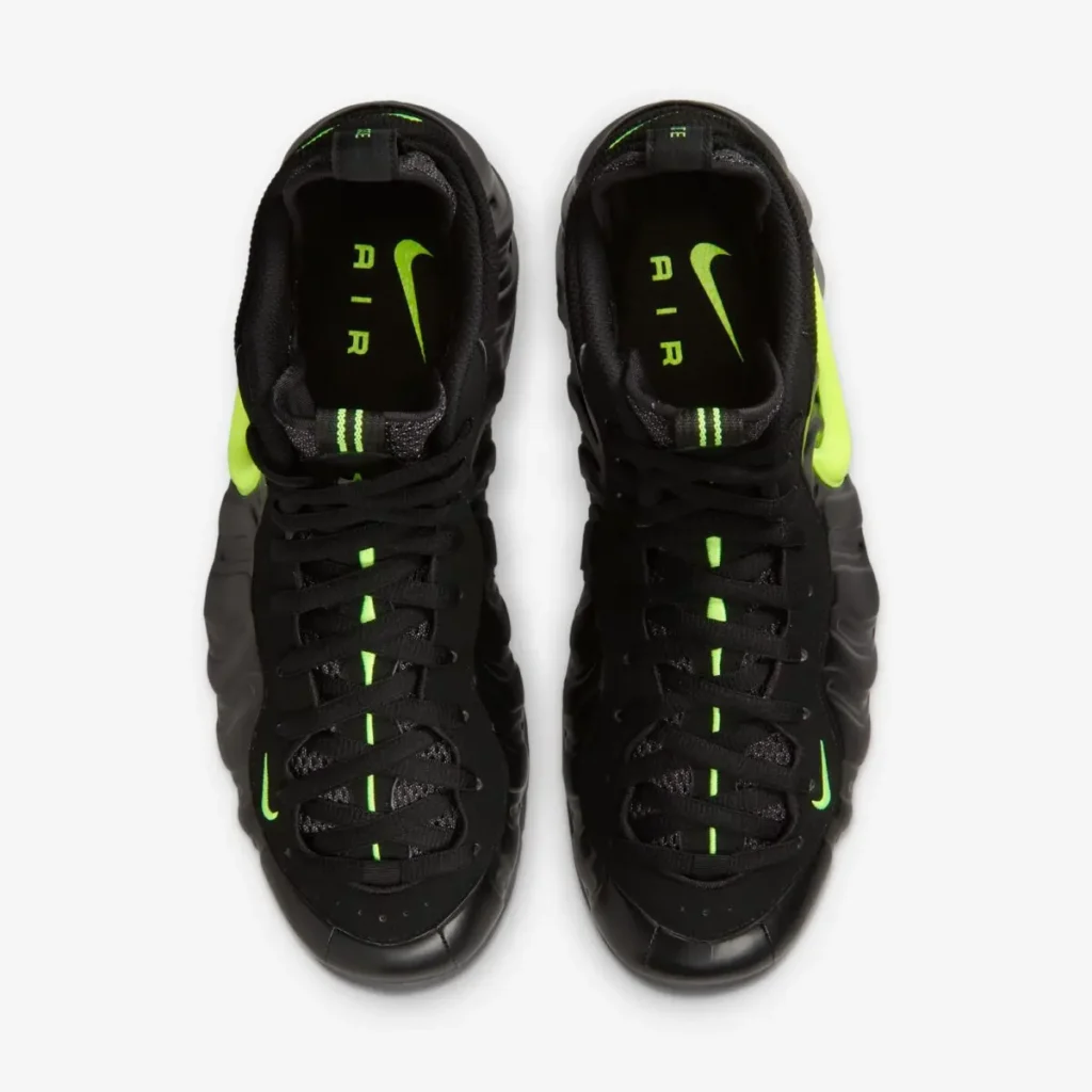 Top-down view of the Nike Air Foamposite Pro “Voltage,” showing black laces over a mesh tongue with a neon Voltage Yellow stripe running down the center, small Swooshes near the toe, heel pull tabs with striped detailing, and insoles featuring a bright green Nike Swoosh with “AIR” lettering