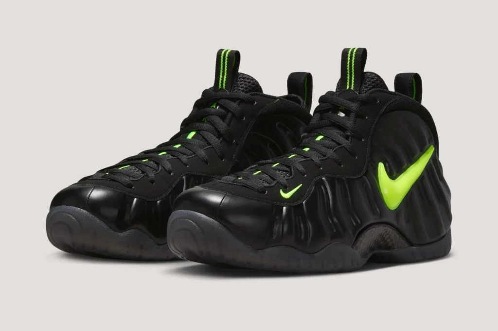 Angled front three-quarter view of the Nike Air Foamposite Pro “Voltage,” showcasing the glossy black molded shell with wave-like ridges, a bold neon Voltage Yellow Swoosh on the lateral side, black laces over a mesh tongue with green accents, striped pull tabs, and a smoky translucent outsole wrapping the sculpted base