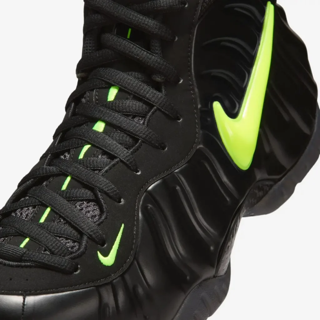 Close-up detail of the Nike Air Foamposite Pro “Voltage,” highlighting the glossy black molded shell with wave-like ridges, a bold neon Voltage Yellow lateral Swoosh, black laces over a mesh tongue with a vertical green stripe, and a small embroidered mini Swoosh near the forefoot