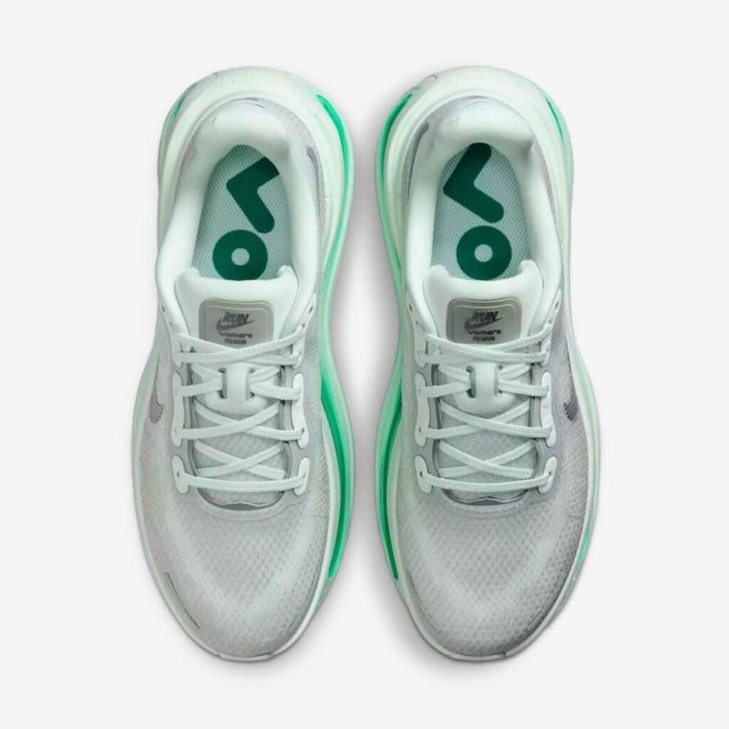 Top-down view of the Nike Vomero Premium Women’s “Barely Green” (HM5973-300), showing the soft green engineered mesh upper, tonal lacing system, padded collar, and insole branding with bold green “ZOOM” lettering, framed by a subtle metallic silver Swoosh and translucent midsole edges
