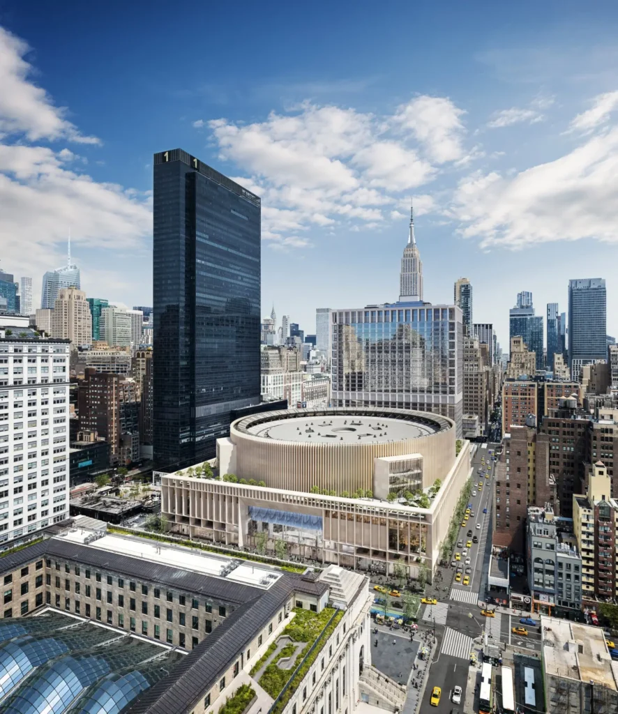 Daytime aerial rendering of a redesigned Penn Station district in Midtown Manhattan, featuring a large circular arena structure rising above a multi-level podium, surrounded by dense high-rise towers with the Empire State Building visible in the skyline under a bright blue sky
