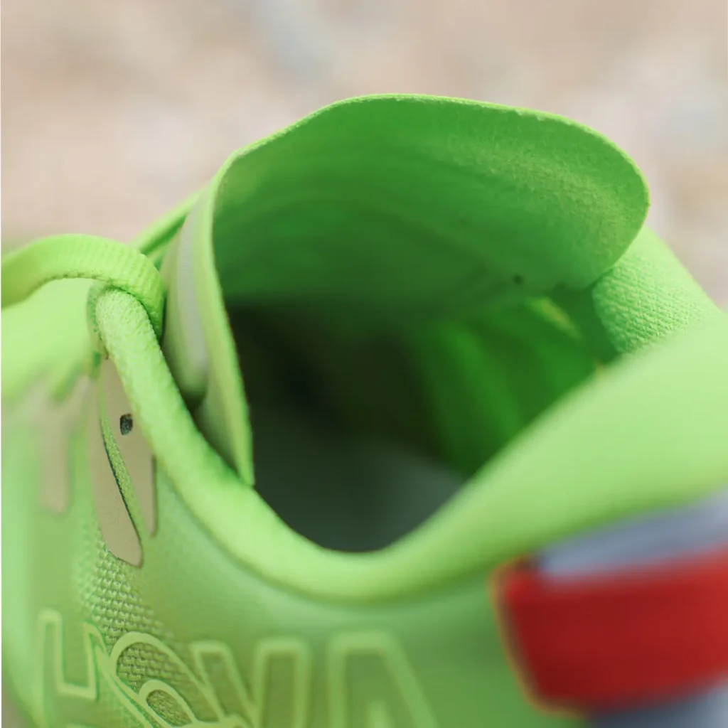 Extreme close-up of a trail running shoe collar and interior opening, focusing on comfort-oriented construction and material detailing. The shoe is finished in a vivid lime green colorway with a padded ankle collar, soft tongue lining, and tonal branding integrated into the side panel. A contrasting red heel pull tab adds a bold accent at the rear, while the shallow depth of field emphasizes cushioning, interior fit, and the lightweight technical build designed for long-distance trail comfort