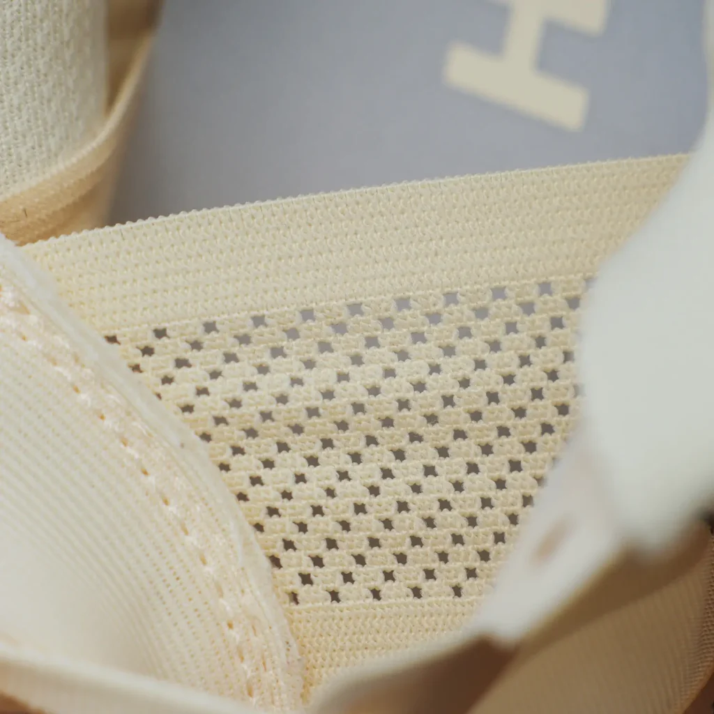 Macro close-up of a trail running shoe interior focusing on the engineered mesh construction and breathable collar design. The image highlights a soft cream-toned woven lining with an open-grid ventilation pattern across the tongue and inner upper, framed by tonal lace and collar elements. The shallow depth of field draws attention to airflow, lightweight comfort, and precision textile detailing, emphasizing the shoe’s performance-oriented interior architecture for extended trail use