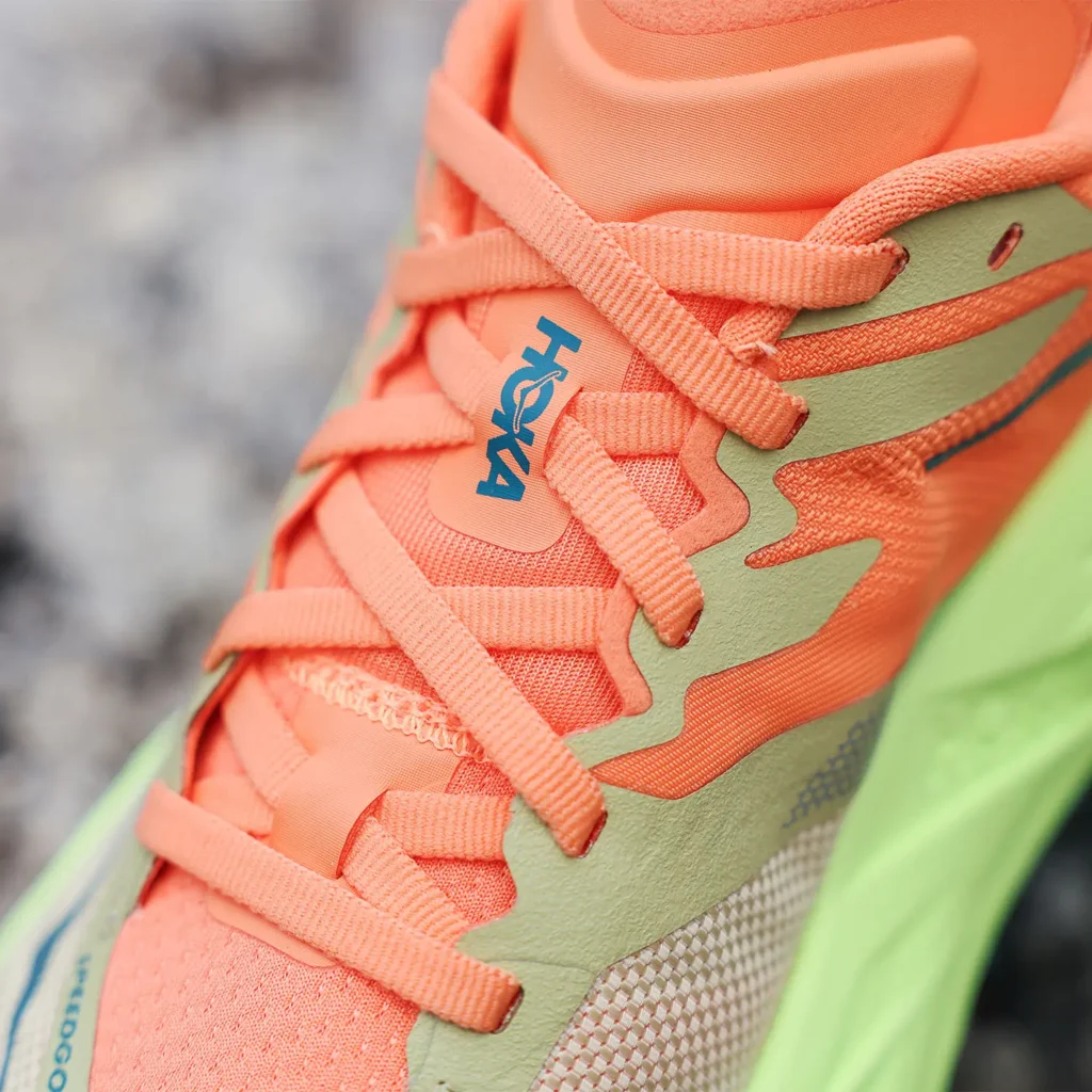 Macro detail of a technical trail running shoe upper focusing on the lace system, branding, and layered material construction. The shoe combines a vivid coral-orange engineered mesh upper with tonal flat laces and olive-green structural overlays that create a dynamic zigzag eyestay pattern. A blue logo mark appears vertically on the tongue, while the bright lime-green sculpted midsole enters the frame at the edge, reinforcing the model’s energetic outdoor palette. The close crop emphasizes lightweight support, breathable textiles, and modern performance-focused design for trail running
