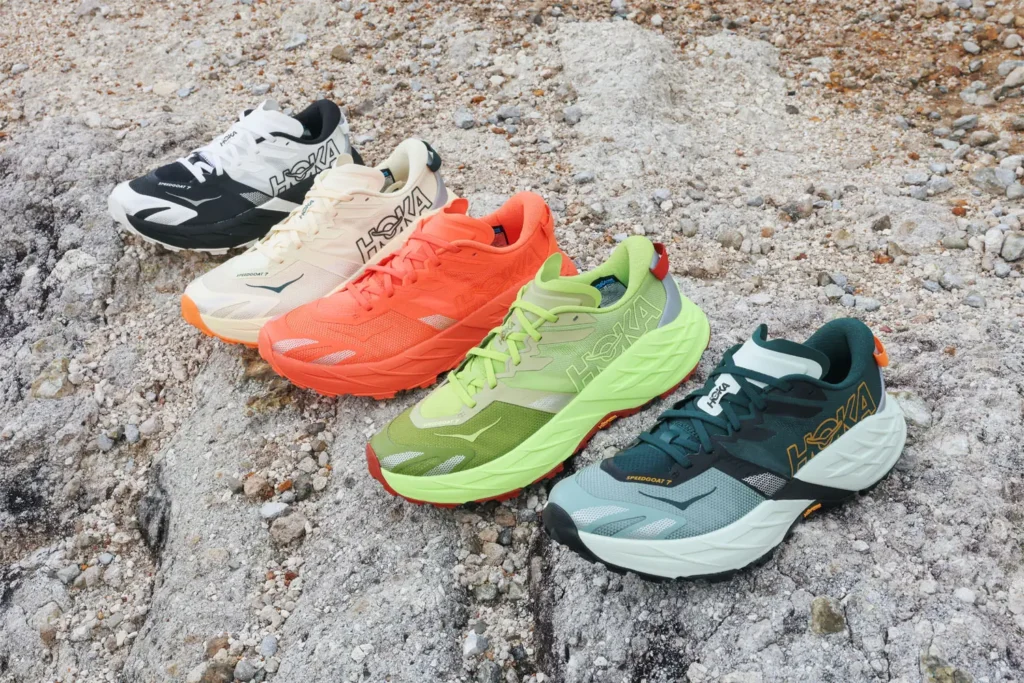 Group display of five trail running shoe colorways arranged across pale rocky terrain, showcasing the model’s range of outdoor-ready finishes and technical construction. The lineup includes black-and-white, cream, vivid coral, lime green, and grey-teal variations, each featuring oversized side branding, breathable engineered mesh uppers, layered support panels, and sculpted high-stack midsoles. Positioned in a staggered formation, the composition highlights differences in color, cushioning geometry, and rugged outsole design while reinforcing the collection’s trail-focused performance identity in a natural outdoor setting