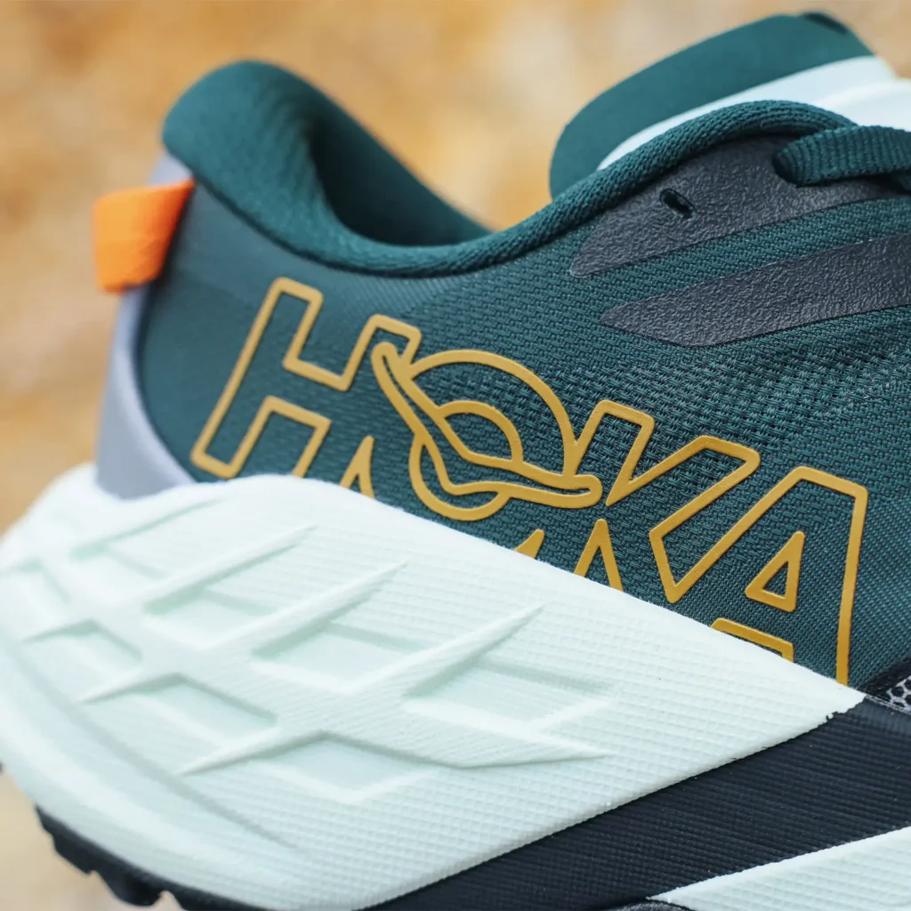 Extreme close-up detail of a technical trail running shoe highlighting the oversized sidewall branding and layered construction. The shoe features a deep teal engineered mesh upper with outlined tonal logo graphics, a sculpted white foam midsole with angular geometric texturing, and subtle orange heel-tab accents. Cropped framing emphasizes material texture, cushioning architecture, and lightweight outdoor performance design while keeping the rocky terrain background softly out of focus