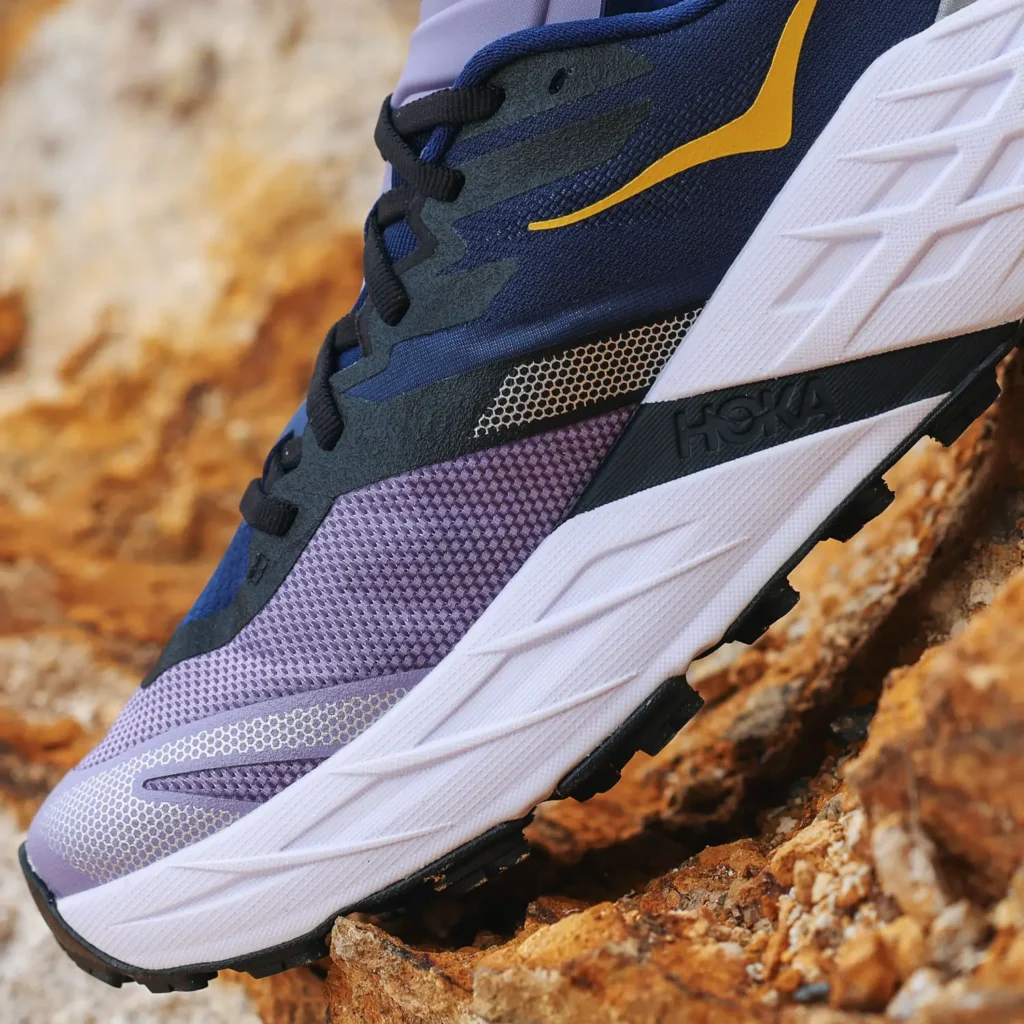 Close-up side profile of a technical trail running shoe positioned against rugged stone terrain, emphasizing layered construction and off-road performance detailing. The shoe combines deep navy and black overlays with a lavender forefoot panel, accented by a sharp yellow side logo and subtle reflective texturing. A thick sculpted white foam midsole with angular geometry sits above an aggressive black lug outsole, highlighting cushioning, stability, and traction designed for technical mountain and trail environments
