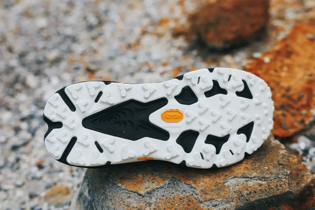 Bottom-outsole detail shot of a trail running shoe resting on rugged rock, highlighting the aggressive traction layout and technical underfoot design. The outsole features a predominantly white rubber construction with deep multidirectional chevron-style lugs, black cutout zones for weight reduction and flexibility, and a contrasting yellow performance rubber badge positioned near the midfoot. The composition emphasizes grip, durability, and terrain control for technical trail and mountain running, with the rocky background softly blurred to keep focus on the tread pattern