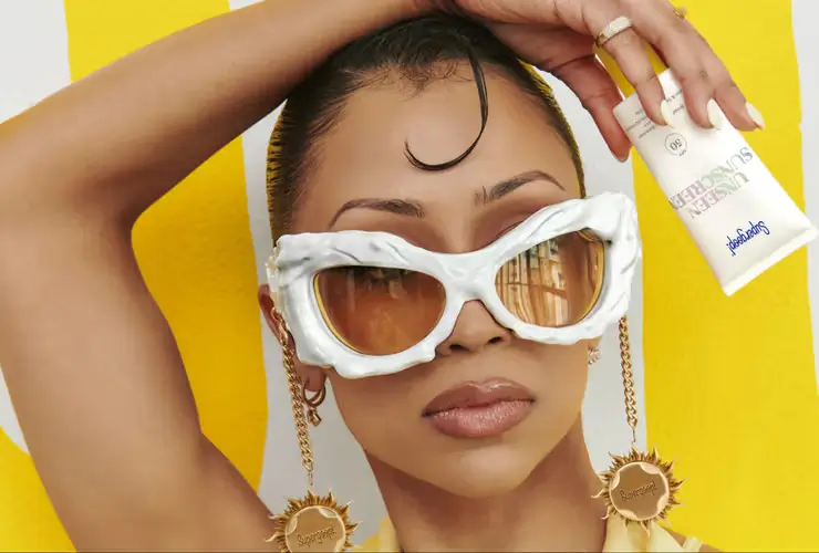 Close-up beauty portrait of a model wearing oversized sculptural white sunglasses with gold-tinted lenses and dangling sun-shaped charms against a bright yellow background
