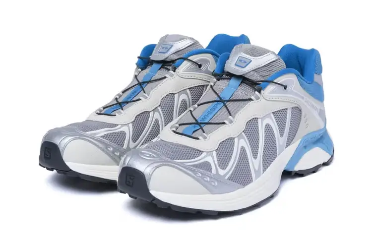 Product image of the SOPHNET. x Salomon XT-Whisper collaboration sneaker in a clean studio setting. The trail-inspired silhouette features silver mesh uppers with metallic overlays, icy blue accents, black quick-lace detailing, and rugged outsole construction, blending technical outdoor performance aesthetics with streamlined contemporary streetwear design