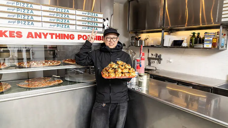 Interior photo of a modern New York-style pizza shop kitchen with a person standing behind the counter holding a tray piled with baked pizza rolls or savory pastries. The person wears glasses, a black cap, and an all-black outfit while gesturing upward with one hand. Behind them, stacks of branded pizza boxes line the wall above a glass display case showcasing whole pizzas and slices. Stainless steel counters, open kitchen equipment, and bright lighting create a clean, industrial atmosphere that blends food culture with a creative, neighborhood hangout aesthetic