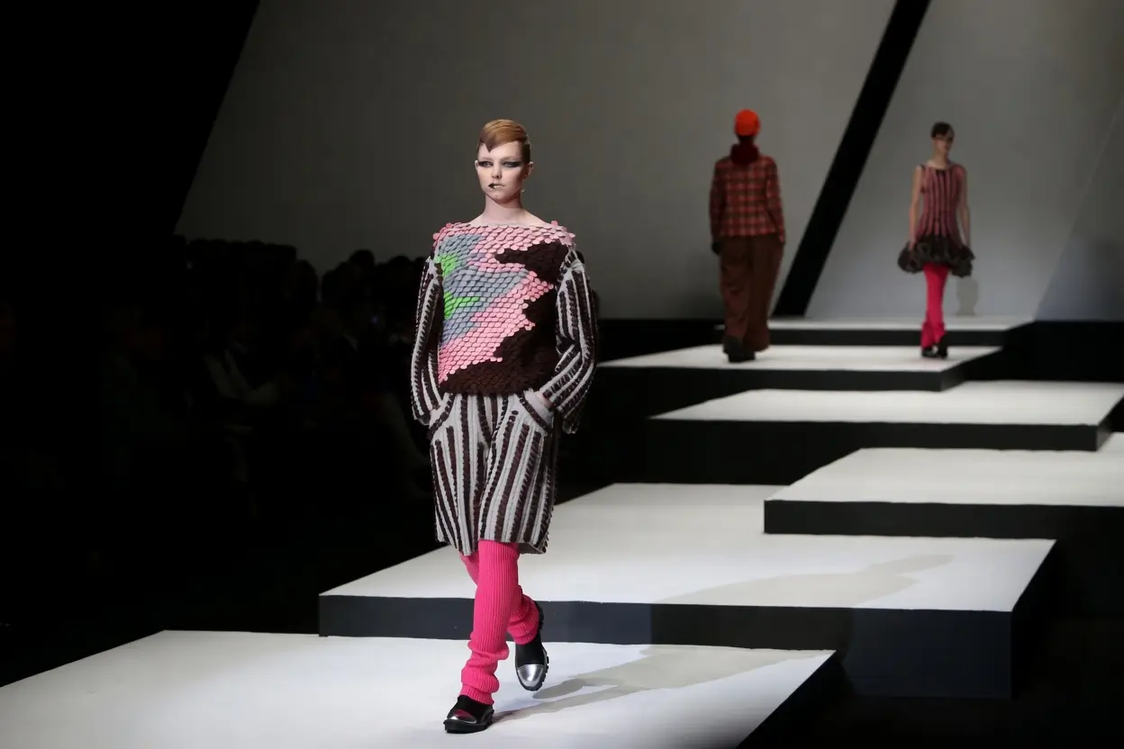 Runway presentation featuring a model walking along a geometric black-and-white catwalk in an avant-garde knit look with sculptural texture, striped proportions, and vivid pink legwear, while additional looks appear in the background—capturing Hiroko Koshino’s signature blend of experimental silhouette, graphic rhythm, and art-driven fashion performance