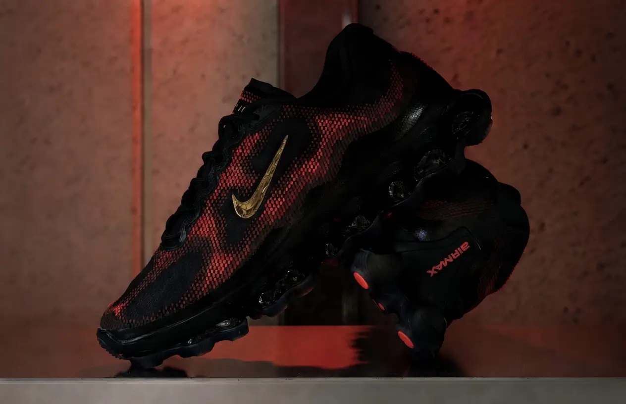 Atmospheric campaign image of the Nike Air Liquid Max “Black/Team Red” suspended in a dramatic studio setup, highlighting the liquid-inspired red-and-black textured upper, metallic gold Swoosh, sculpted Air cushioning pods, and glowing red outsole details against moody industrial lighting
