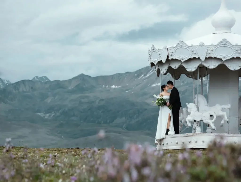 A bride and groom embrace inside an ornate white carousel pavilion positioned in a vast alpine meadow, with muted mountain ranges and cloudy skies creating a cinematic pre-wedding landscape scene