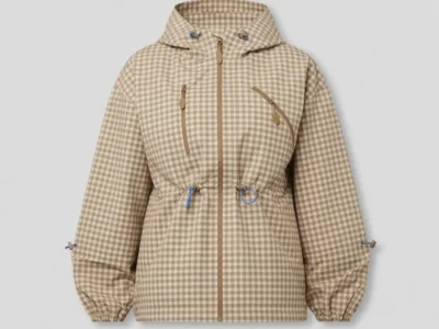 A front-facing studio image of a hooded technical jacket covered in a soft beige-and-cream gingham pattern. The silhouette combines utilitarian outdoor detailing with a refined, fashion-oriented finish, featuring a full front zip closure, angled chest zip pockets, and adjustable drawcords at the waist, hood, and sleeves. Bright blue cord accents subtly contrast against the otherwise muted palette, while elasticated cuffs and structured seam placement reinforce the garment’s lightweight functional design. Set against a clean gray backdrop, the piece balances countryside nostalgia with contemporary outerwear sensibilities