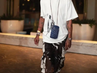 Travis Scott steps through a luxury hotel interior wearing the Air Jordan 1 Low OG “Pink Pack,” styled with oversized graphic layers, patterned shorts, and a Chanel crossbody bag