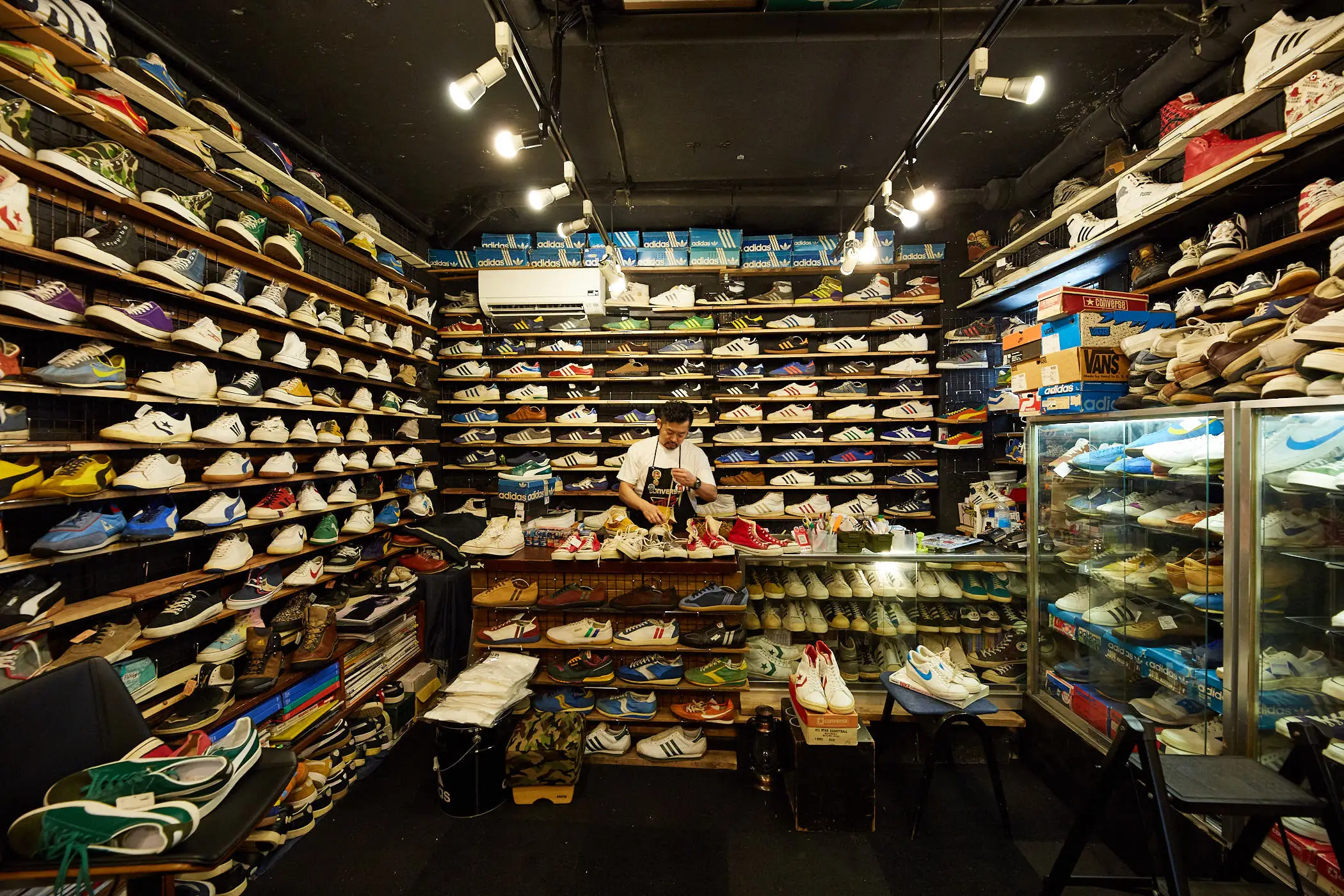 A densely packed vintage sneaker boutique filled wall-to-wall with archival Adidas, Vans, Converse, and Nike footwear, where a shopkeeper organizes pairs beneath warm industrial lighting surrounded by stacked shoeboxes and glass display cases