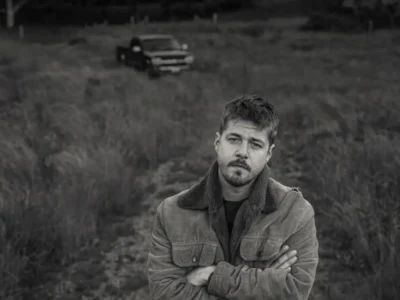 A moody black-and-white portrait of a country musician standing in a field with arms crossed, wearing a shearling-collar jacket while an old pickup truck sits blurred in the background, creating a reflective Americana atmosphere