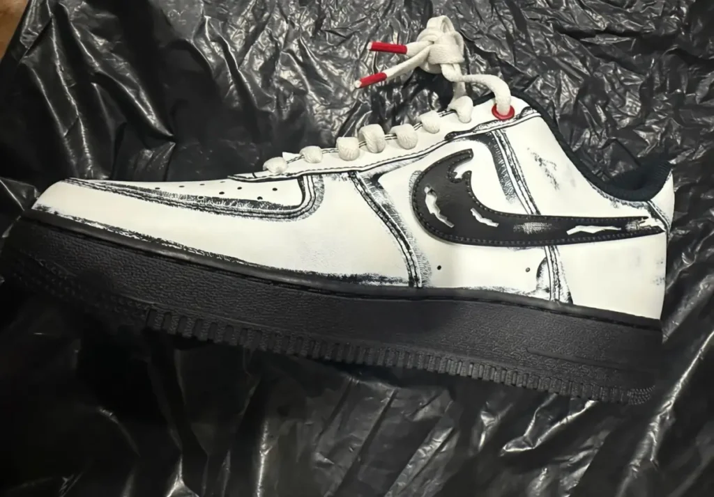 A side-profile view of a customized Nike low-top sneaker showcasing a bold sketch-inspired aesthetic. The shoe features a white leather upper outlined with rough black hand-drawn detailing that mimics illustrated comic-book strokes, giving the silhouette an intentionally unfinished, artistic appearance. A distressed black Swoosh stretches across the side panel, while subtle scuff-like textures and exaggerated seam lines reinforce the handmade visual identity. The sneaker is paired with oversized cream rope laces finished with vivid red aglets, adding contrast against the monochrome design. A fully black midsole and outsole ground the composition with a heavier, more aggressive look, balancing the bright upper with a darker foundation. Set against a wrinkled black plastic backdrop, the image carries a raw prototype energy that blends streetwear customization culture with experimental graphic footwear design